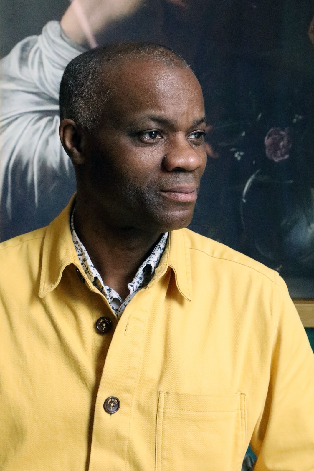 Portrait photograph of Leslie Primo in a yellow jacket, looking to one side, in front of a painting.